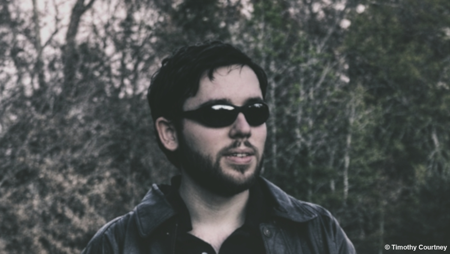 Timothy Courtney is standing near woods wearing a leather jacket like a werewolf